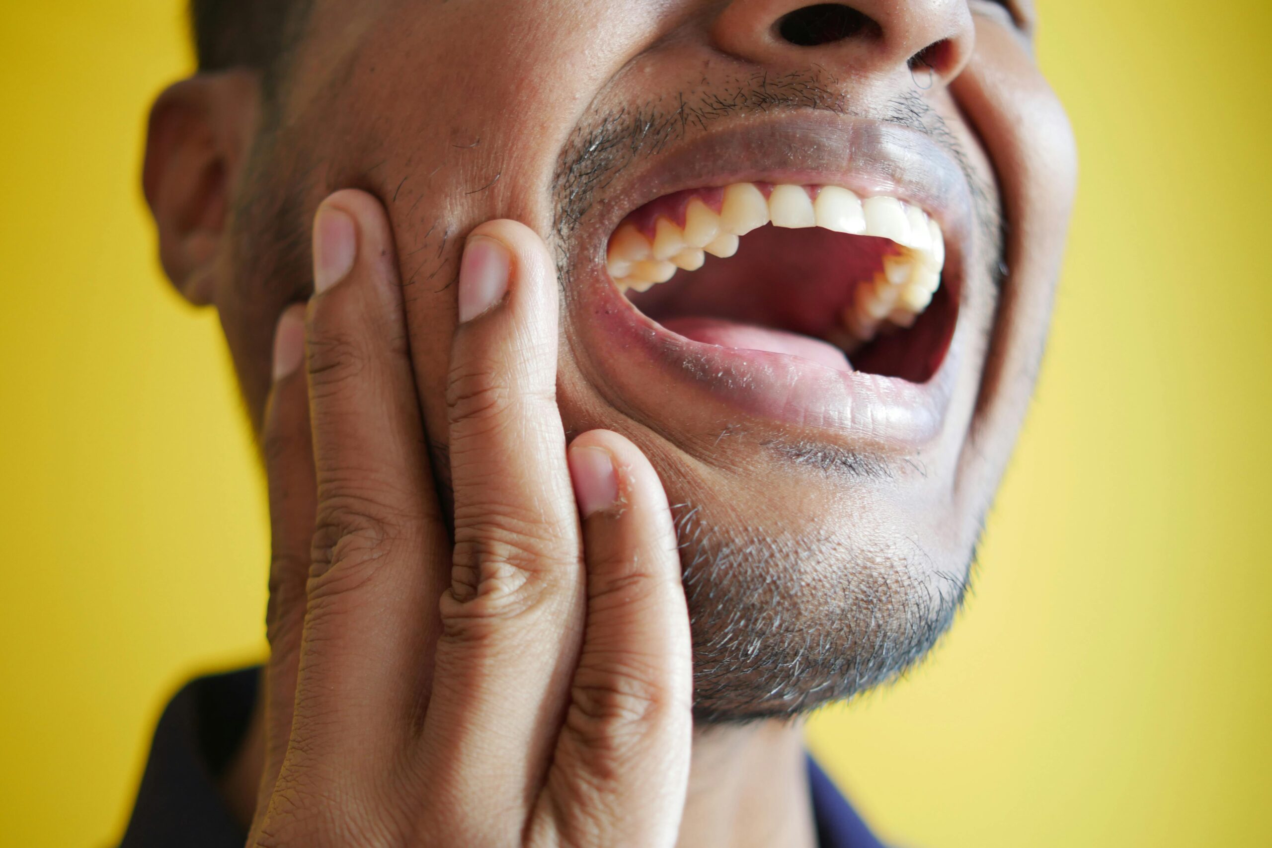 Close-up of a man holding his cheek, displaying a wide open mouth against a yellow background.